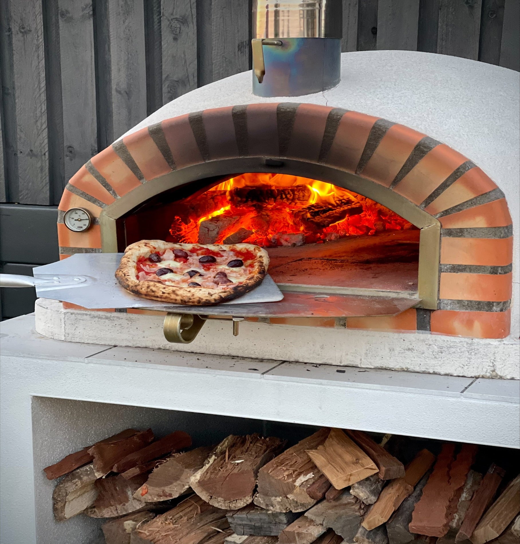 Wood-fired pizza oven with a pizza inside, surrounded by firewood.