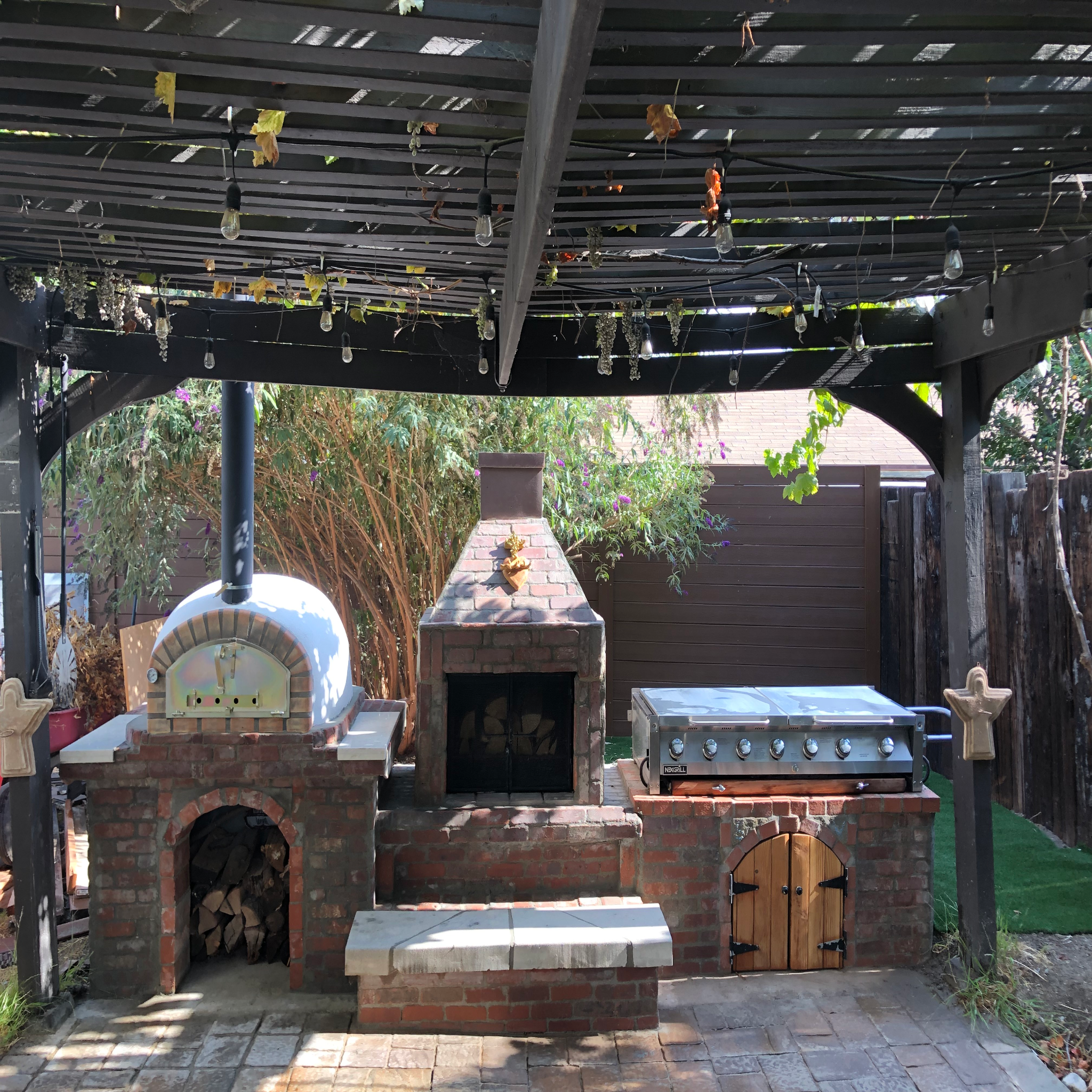Outdoor kitchen with brick oven, grill, and stone bench under a wooden pergola.